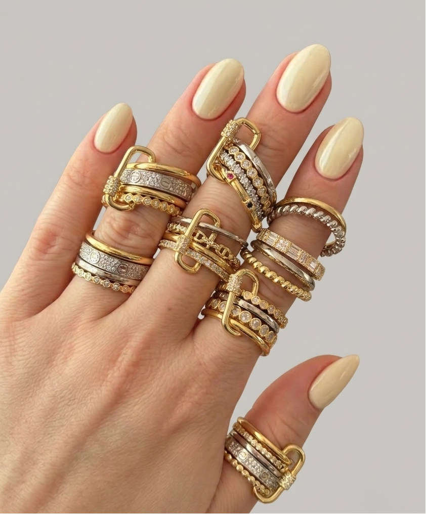 A hand with cream-colored manicured nails wearing an assortment of gold and silver multi-band rings, set against a solid gray background.